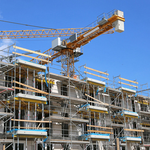 Eine Baustelle für ein mehrstöckiges Gebäude im Rohbau, umgeben von einem Gerüst. Im Hintergrund steht ein großer Baukran unter klarem, blauem Himmel.