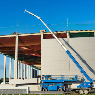 Eine Baustelle für ein Industriegebäude mit Stahlbetonskelett und hohen Wänden. Ein blauer Mobilkran hebt Bauelemente an die Struktur, während Sicherheitsnetze über den offenen Bereichen gespannt sind.