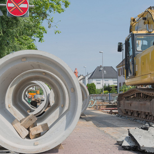 Gelber Bagger steht vor einer offenen Straße für Bauarbeiten. Daneben liegt Baumaterial für den Tiefbau.