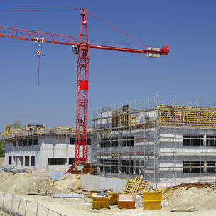 Eine Baustelle für ein mehrstöckiges Gebäude im Rohbau, umgeben von einem Gerüst. Daneben steht ein großer roter Baukran unter klarem, blauem Himmel.