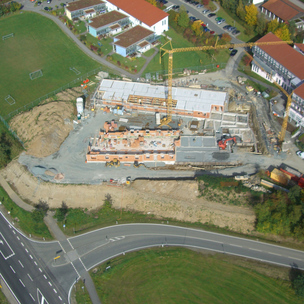 Luftaufnahme einer Baustelle neben einer Straße und einem Sportplatz. Ein Kran und mehrere Baumaschinen sind auf dem Gelände im Einsatz. Gebäude im Rohbau mit erkennbaren Grundrissen und teilweise errichteten Mauern.